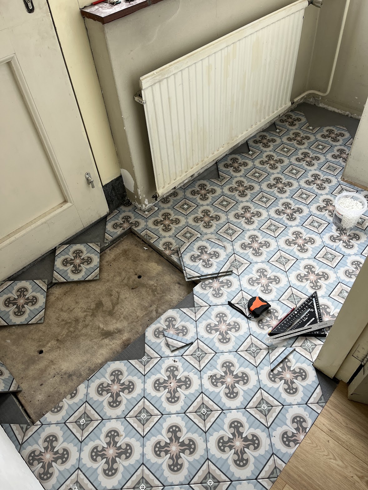 Patterned floor tiles being laid around a radiator, door and unfinished floor section