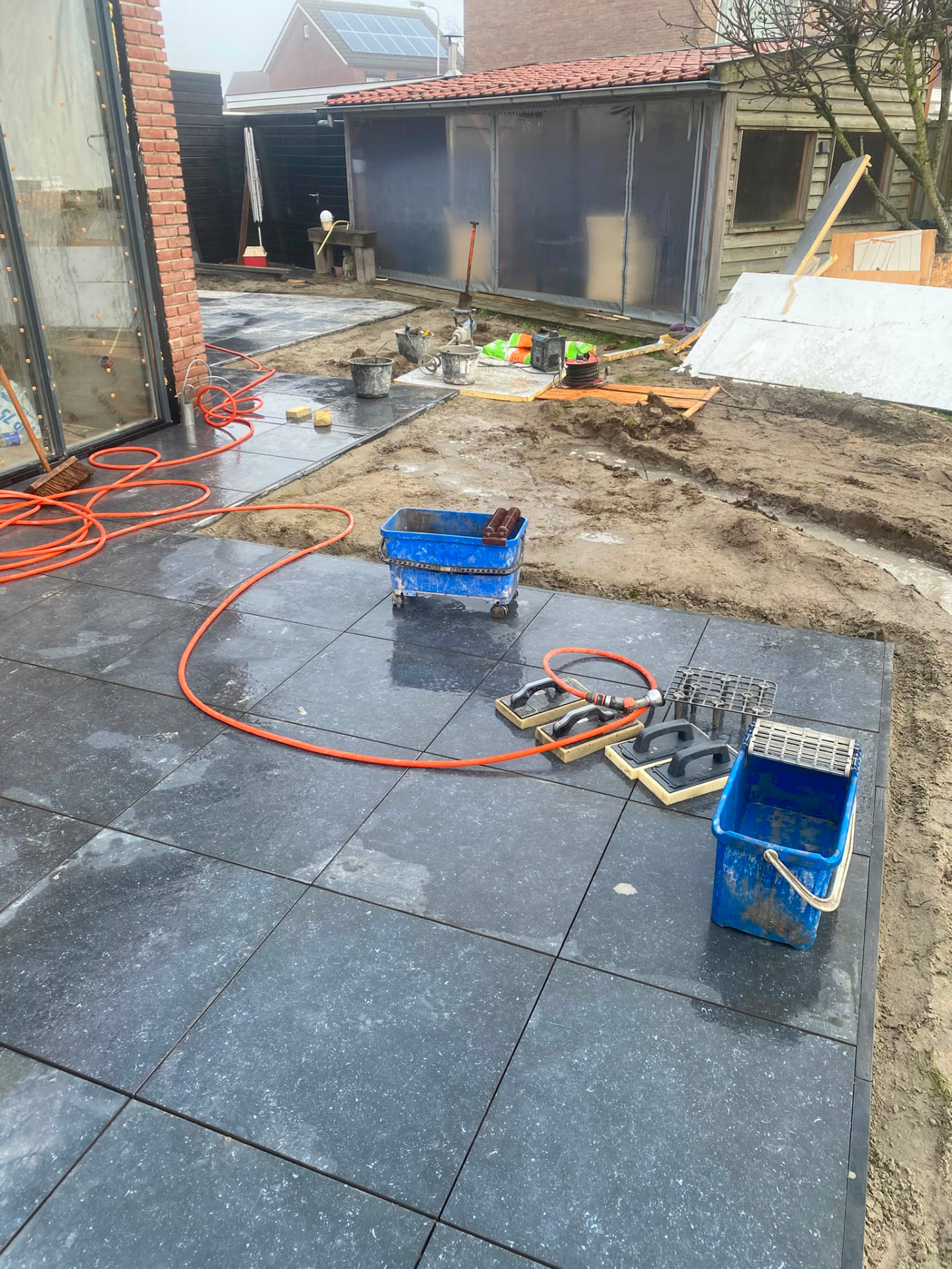 Partially completed outdoor patio with dark square tiles, tools and unfinished soil
