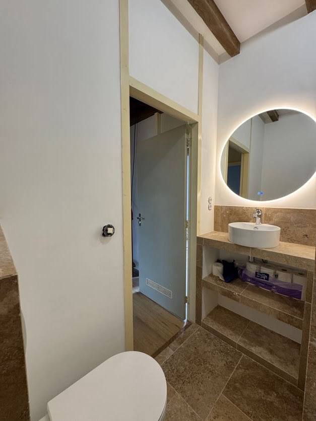 Beige tiled bathroom with round illuminated mirror, stone-effect vanity, open shelving and toilet