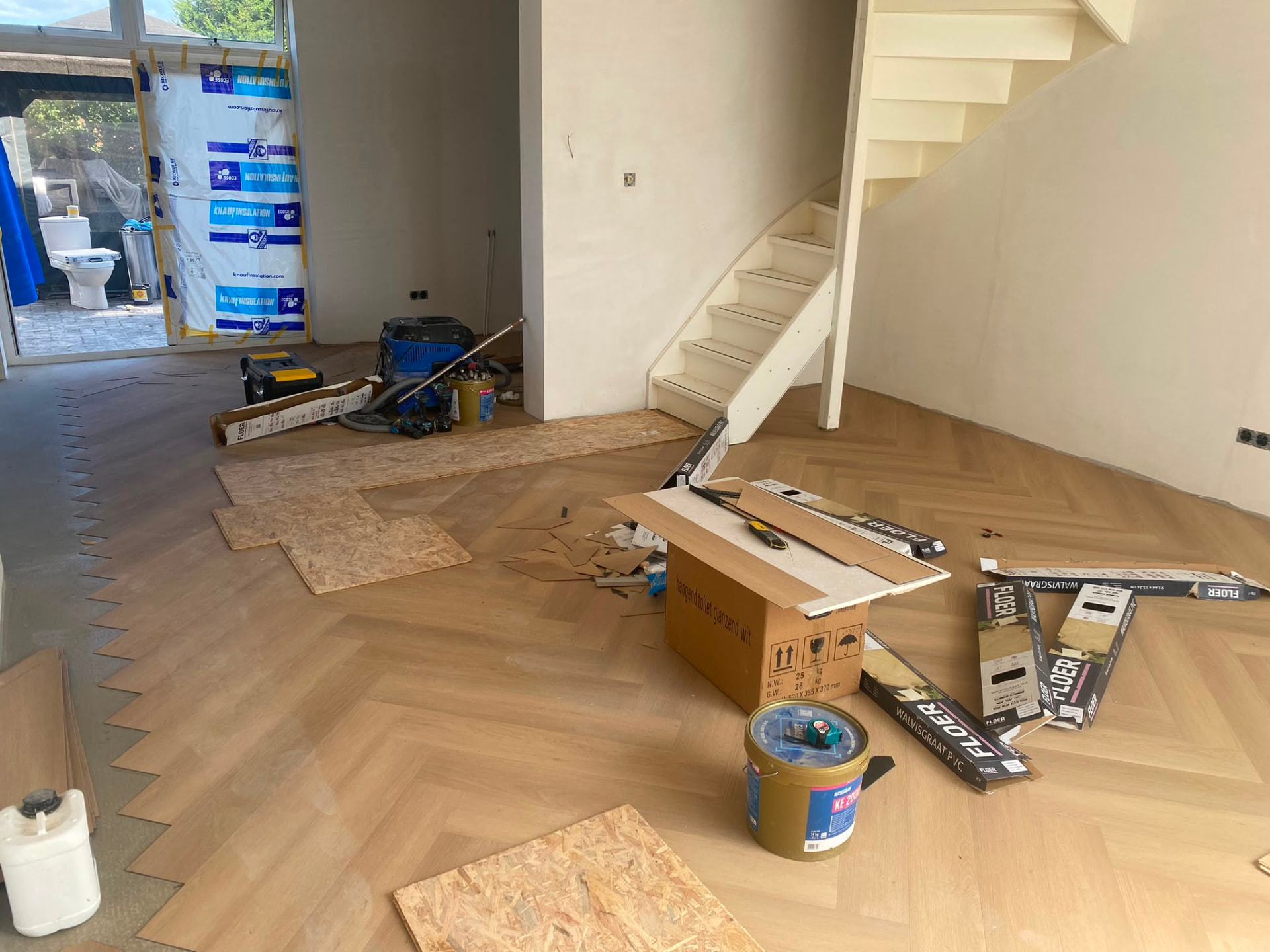 Herringbone laminate flooring being installed at the base of a modern staircase with tools